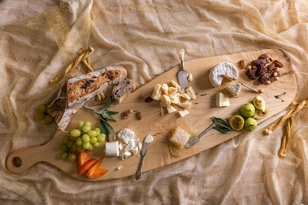 Wooden cutting board with assorted cheeses, fruits, and bread on a textured fabric background